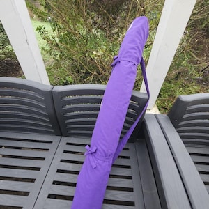 May include: A long, cylindrical purple yoga mat bag with a shoulder strap and a bow closure. The bag is made of a solid, textured fabric and is designed to carry a rolled-up yoga mat. The bag is resting on a gray bench.