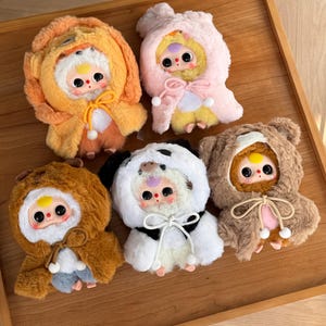 May include: A collection of plush dolls, each dressed in a different animal-themed costume. The costumes include a lion, a bear, a panda, and others, all with soft, fluffy textures and cute facial features. The dolls are arranged on a wooden tray.