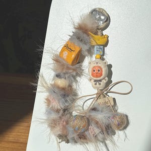 May include: A whimsical keychain featuring a fluffy, multi-colored feather accent. It includes a small orange coffee box charm, a yellow banana, and a cute bear-shaped figure. The keychain is adorned with small decorative balls and a tag that says "Made".