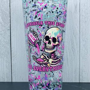 May include: A clear tumbler with a screw-on lid filled with colorful glitter and sprinkles. The tumbler features a cartoon skeleton graphic with the words "Sprinkle this stuff on everything" and "Sarcasm".