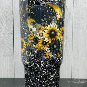 May include: A clear tumbler with a black glitter base and a floral design. The design features sunflowers, a crescent moon, and a butterfly. The tumbler has a clear handle and lid. The overall theme is celestial and floral.