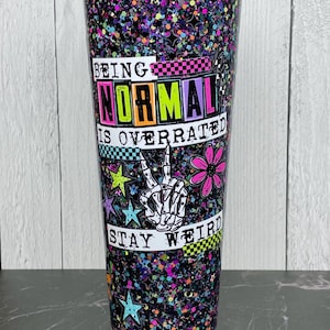 May include: A tall, glitter-covered tumbler with a clear lid. The tumbler features the text "BEING NORMAL IS OVERRATED STAY WEIRD" with a peace sign hand gesture, stars, and a flower graphic. The tumbler is black with colorful glitter.
