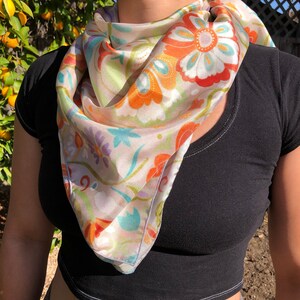 May include: A white silk scarf with a floral pattern in shades of orange, blue, green, and purple. The scarf is tied around the neck of a person wearing a black shirt.