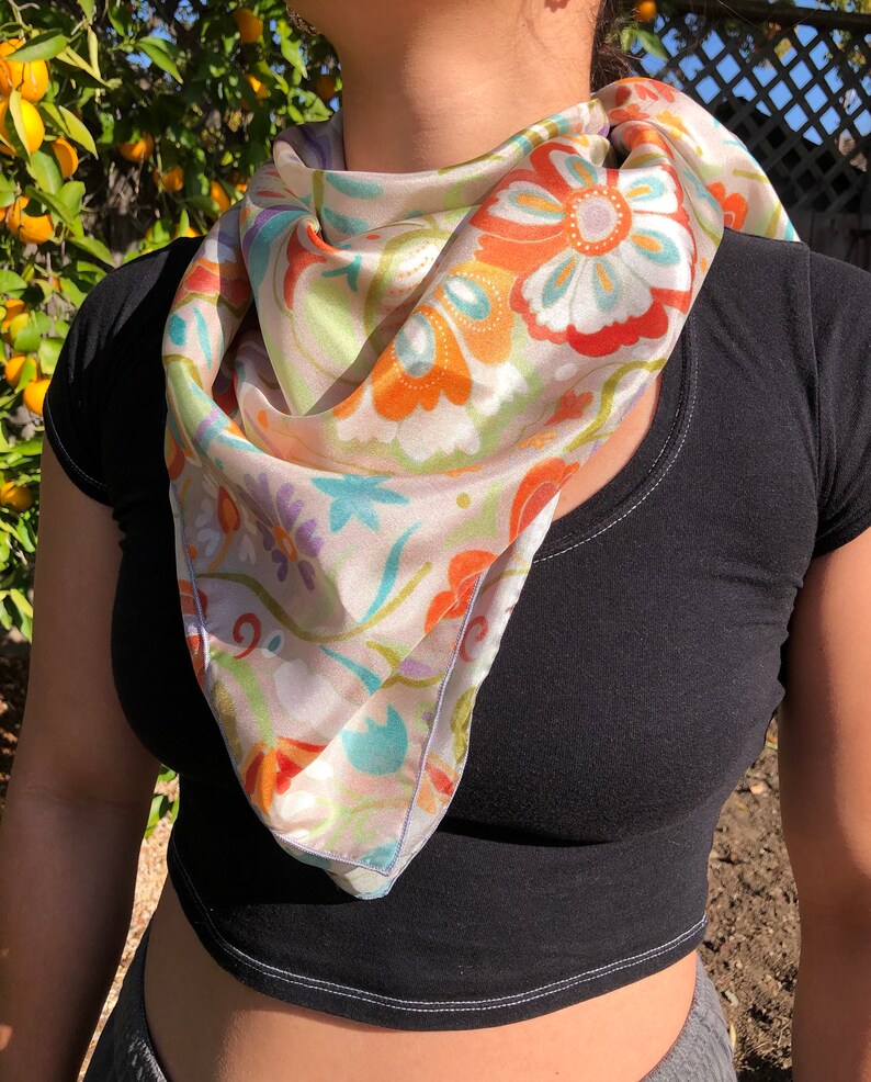 May include: A white silk scarf with a floral pattern in shades of orange, blue, green, and purple. The scarf is tied around the neck of a person wearing a black shirt.