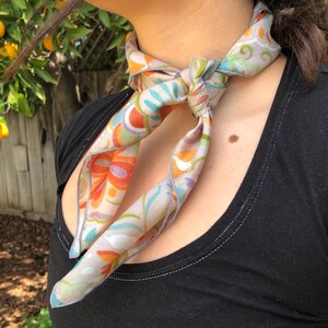 May include: A gray silk scarf with a floral pattern in shades of orange, blue, and green. The scarf is tied around the neck in a knot.