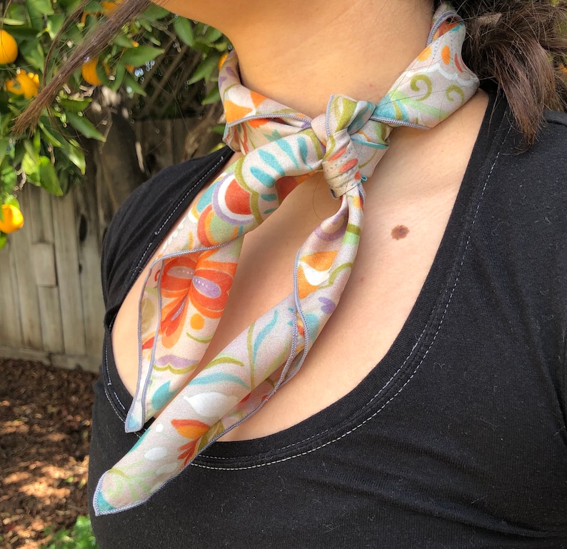May include: A gray silk scarf with a floral pattern in shades of orange, blue, and green. The scarf is tied around the neck in a knot.