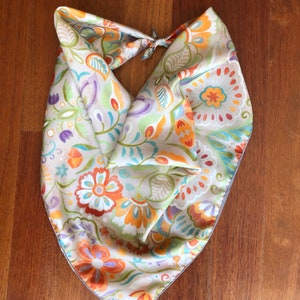 May include: A beige silk scarf with a floral pattern in shades of orange, green, purple, and blue. The scarf is tied in a knot at the top.