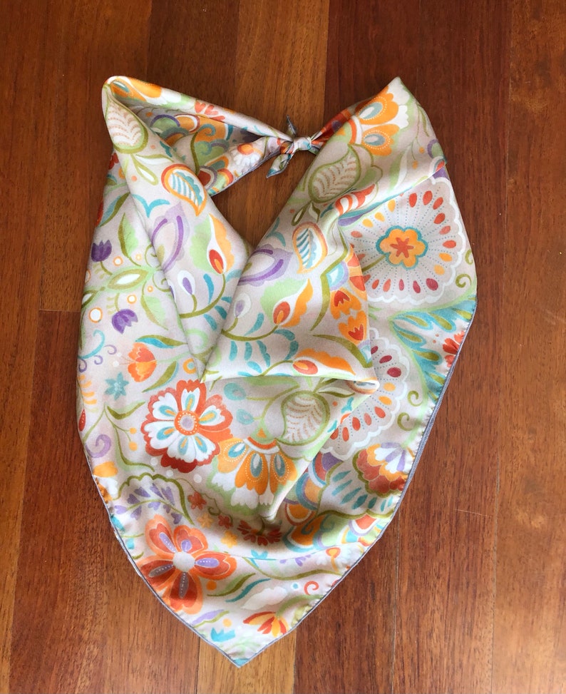 May include: A beige silk scarf with a floral pattern in shades of orange, green, purple, and blue. The scarf is tied in a knot at the top.
