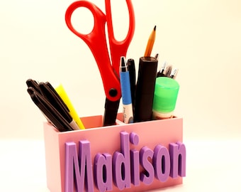 May include: Pink desk organiser with the name "Madison" printed on the front. The organiser has three compartments and is filled with various school supplies, including scissors, pens, pencils, and a glue stick.