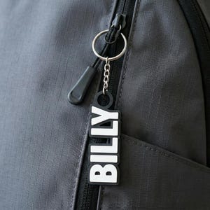May include: A black and white keychain with the name "BILLY" in bold, capital letters hangs from a silver chain attached to a black zip. The keychain is against a dark grey backpack.