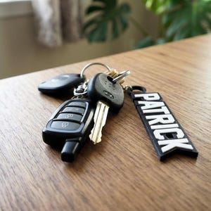 May include: A set of car keys and a key fob with the name "PATRICK" in white lettering on a black background. The keys are on a silver key ring and are resting on a wooden surface.