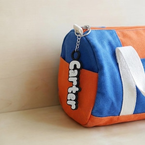May include: A blue and orange duffel bag with a white strap and a black and white keychain that says "Carter". The bag is made of canvas material and has a zip closure.