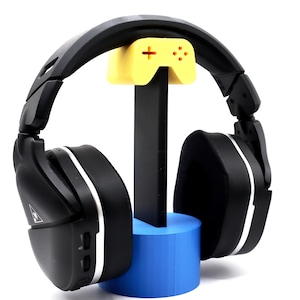 Peut inclure: Casque de jeu noir avec un accent argenté sur les écouteurs, reposant sur un support bleu et jaune. Le support a un dessus en forme de manette.