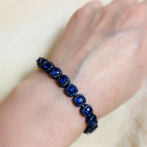 Pu&ograve; includere: Un braccialetto blu scuro con pietre preziose quadrate e scintillanti. Il braccialetto &egrave; indossato su un polso, mettendo in risalto il design intricato e il colore vibrante del gioiello. Le pietre sono incastonate vicine, creando una fascia continua di blu.