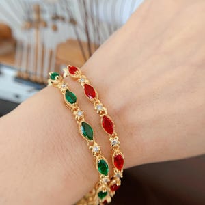 May include: Two gold-toned bracelets with alternating red and green gemstone accents. Each bracelet features a series of marquise-cut gemstones and small, clear crystals. The bracelets are displayed on a person's wrist.