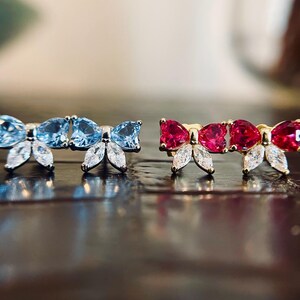 May include: Two pairs of bow-shaped stud earrings. One pair features light blue gemstones and white accents, while the other showcases red gemstones and white accents. The earrings are set on a gold-colored base, reflecting light.