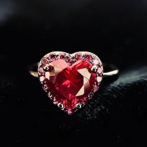 May include: A heart-shaped ring with a large, faceted red gemstone as the centerpiece. The gemstone is surrounded by smaller, pink gemstones. The ring's band is a light silver color, and the background is black.