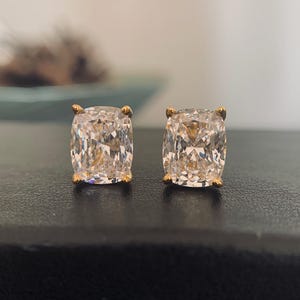 May include: A pair of cushion-cut stud earrings. Each earring features a large, clear, sparkling gemstone set in a gold-colored metal. The earrings are displayed on a dark surface, with a blurred background.