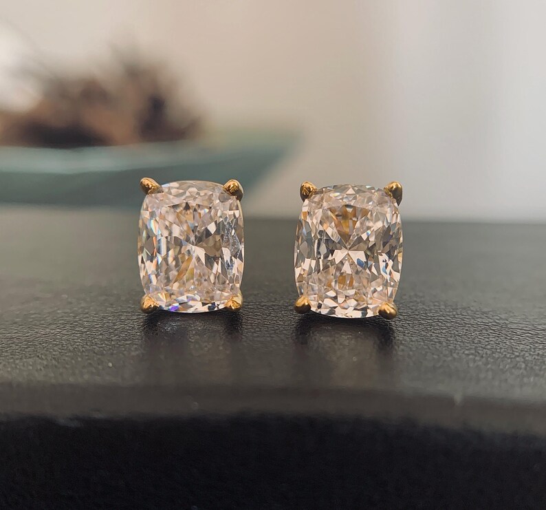 May include: A pair of cushion-cut stud earrings. Each earring features a large, clear, sparkling gemstone set in a gold-colored metal. The earrings are displayed on a dark surface, with a blurred background.
