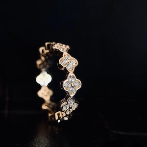 May include: Rose gold ring featuring a repeating clover design. Each clover is adorned with small, sparkling gemstones. The ring is set against a dark background, highlighting its intricate details and elegant design. A beautiful piece of jewelry.