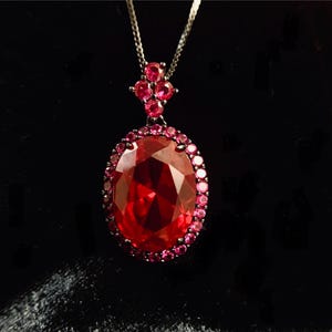 May include: A silver necklace with a large, oval, faceted red gemstone pendant. The pendant is surrounded by a halo of smaller pink gemstones. A cluster of pink gemstones adorns the top of the pendant, and the necklace has a delicate chain.
