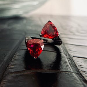 May include: A black ring featuring two triangular, vibrant red gemstones. The gemstones are set in a dark, textured metal band. The ring's design is modern and eye-catching, perfect for adding a pop of color.
