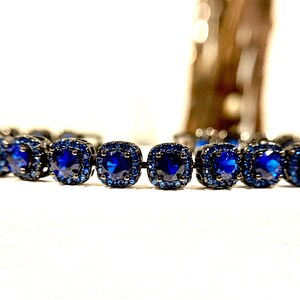 Pu&ograve; includere: Primo piano di un braccialetto con pietre preziose blu scuro a taglio quadrato incastonate in metallo scuro. Le pietre centrali sono circondate da pietre blu pi&ugrave; piccole. Il braccialetto &egrave; appoggiato su una superficie bianca.