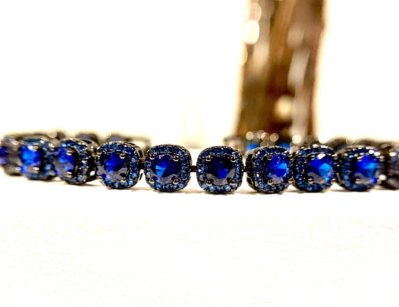 Pu&ograve; includere: Primo piano di un braccialetto con pietre preziose blu scuro a taglio quadrato incastonate in metallo scuro. Le pietre centrali sono circondate da pietre blu pi&ugrave; piccole. Il braccialetto &egrave; appoggiato su una superficie bianca.