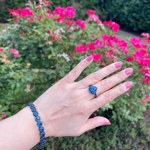 Pu&ograve; includere: Una mano che indossa un anello e un braccialetto con pietre preziose blu. L'anello presenta una pietra blu a forma di cuore incastonata in metallo scuro. Il braccialetto ha pietre blu quadrate. Lo sfondo include fiori rosa e fogliame verde.