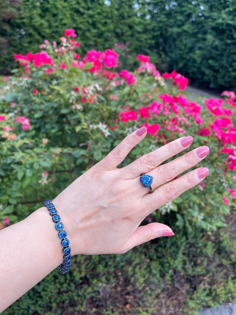 Pu&ograve; includere: Una mano che indossa un anello e un braccialetto con pietre preziose blu. L'anello presenta una pietra blu a forma di cuore incastonata in metallo scuro. Il braccialetto ha pietre blu quadrate. Lo sfondo include fiori rosa e fogliame verde.