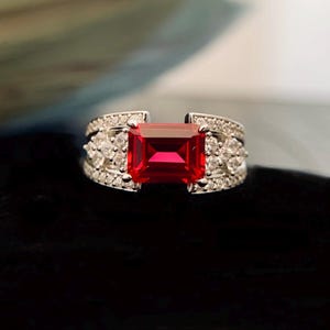 May include: A silver ring with a large, rectangular, bright red gemstone as the focal point. The band is encrusted with smaller, clear gemstones. The ring is set against a dark background, highlighting the sparkle of the gems.