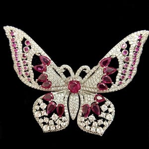 May include: A silver-toned butterfly brooch with a symmetrical design. The wings are encrusted with small, clear crystals and larger, teardrop-shaped, and round ruby-colored stones. The brooch is set against a black background.