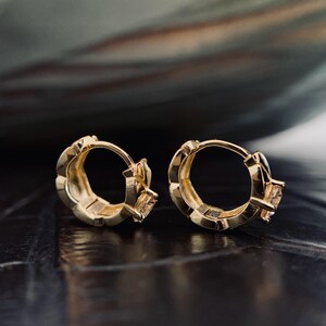 Op de afbeelding: Een paar goudkleurige oorringen. Elke oorring heeft een vierkante edelsteen en een gestructureerd ontwerp langs de ring. De oorbellen worden gepresenteerd op een donker, reflecterend oppervlak.