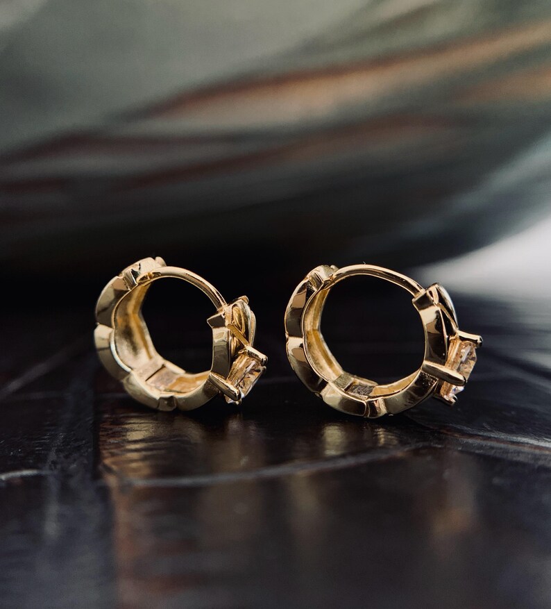 Op de afbeelding: Een paar goudkleurige oorringen. Elke oorring heeft een vierkante edelsteen en een gestructureerd ontwerp langs de ring. De oorbellen worden gepresenteerd op een donker, reflecterend oppervlak.