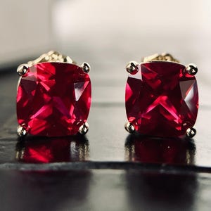 May include: A pair of square-cut, ruby-red gemstone stud earrings. Each earring features a vibrant, faceted gemstone set in a gold-toned metal setting with four small, silver-colored accents. The earrings are displayed on a dark surface.