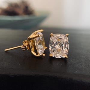 May include: A pair of gold-colored stud earrings with large, square-cut, clear gemstones. The earrings are set in a four-prong setting, and the posts are visible. The background is a dark surface.