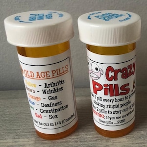 May include: Two orange prescription bottles with white lids. One bottle reads "OLD AGE PILLS" with a list of ailments. The other bottle reads "Crazy Pills" with a warning label. Both bottles have white labels with black and red text.