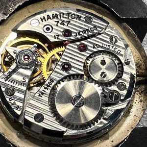 K&ouml;nnte beinhalten: Nahaufnahme eines Vintage Hamilton 747 Uhrwerks. Der Mechanismus verf&uuml;gt &uuml;ber komplizierte Zahnr&auml;der, Juwelen und den Text "HAMILTON 747 JEWELS ADJUSTED U.S.A." Das Uhrwerk ist silber- und goldfarben.