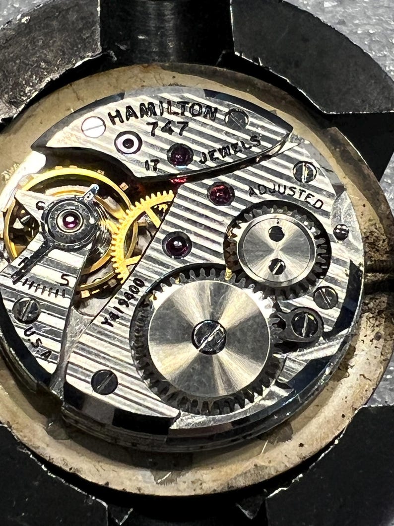 K&ouml;nnte beinhalten: Nahaufnahme eines Vintage Hamilton 747 Uhrwerks. Der Mechanismus verf&uuml;gt &uuml;ber komplizierte Zahnr&auml;der, Juwelen und den Text "HAMILTON 747 JEWELS ADJUSTED U.S.A." Das Uhrwerk ist silber- und goldfarben.