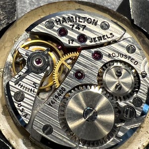 K&ouml;nnte beinhalten: Nahaufnahme eines Vintage Hamilton 747 Uhrwerks. Der silberfarbene Mechanismus verf&uuml;gt &uuml;ber komplizierte Zahnr&auml;der, Rubinjuwelen und den Text "HAMILTON 747 17 JEWELS ADJUSTED U.S.A." Das Uhrwerk ist teilweise zerlegt.