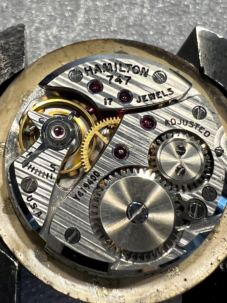 K&ouml;nnte beinhalten: Nahaufnahme eines Vintage Hamilton 747 Uhrwerks. Der silberfarbene Mechanismus verf&uuml;gt &uuml;ber komplizierte Zahnr&auml;der, Rubinjuwelen und den Text "HAMILTON 747 17 JEWELS ADJUSTED U.S.A." Das Uhrwerk ist teilweise zerlegt.