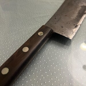 May include: A well-used, vintage cleaver with a dark wooden handle secured by three silver rivets. The rectangular blade shows signs of wear and a small hole near the top. The cleaver is designed for chopping and cutting.