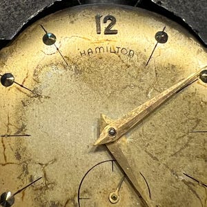 K&ouml;nnte beinhalten: Nahaufnahme eines Vintage-Zifferblatts der Marke Hamilton. Das Zifferblatt hat eine verblasste Goldfarbe mit schwarzen Markierungen und Zeigern. Die Zahl 12 ist prominent oben dargestellt. Das Wort "HAMILTON" ist unter der Zahl 12 aufgedruckt.