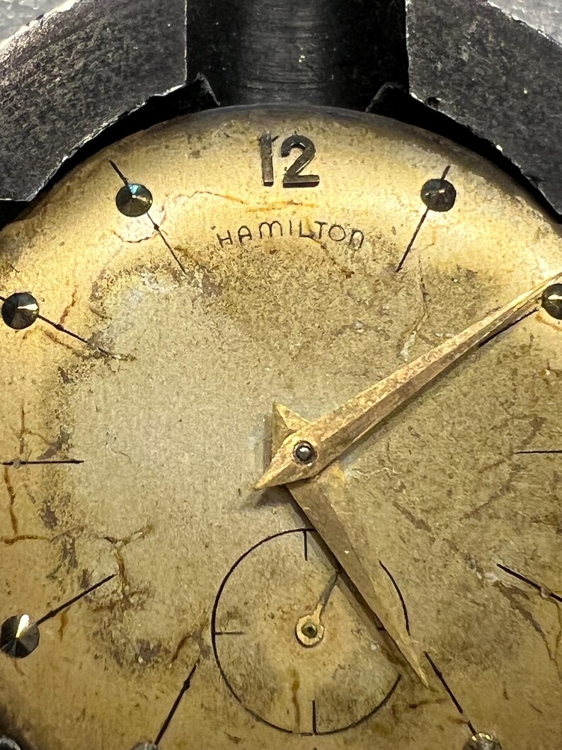 K&ouml;nnte beinhalten: Nahaufnahme eines Vintage-Zifferblatts der Marke Hamilton. Das Zifferblatt hat eine verblasste Goldfarbe mit schwarzen Markierungen und Zeigern. Die Zahl 12 ist prominent oben dargestellt. Das Wort "HAMILTON" ist unter der Zahl 12 aufgedruckt.