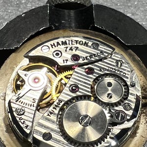 K&ouml;nnte beinhalten: Nahaufnahme eines Vintage Hamilton 747 Uhrwerks. Der komplizierte Mechanismus zeigt goldene Zahnr&auml;der, silberne Komponenten und Rubinjuwelen. Der Text "HAMILTON 747 17 JEWELS ADJUSTED USA" ist sichtbar. Die Uhr wird in einer Metallklemme gehalten.
