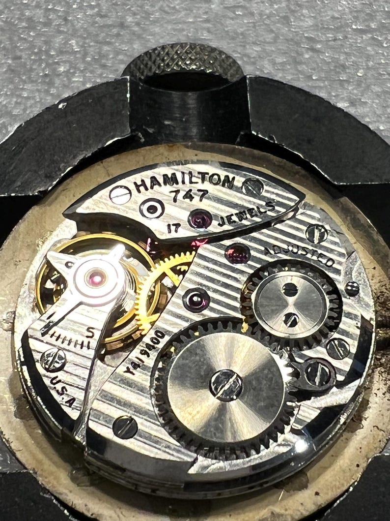 K&ouml;nnte beinhalten: Nahaufnahme eines Vintage Hamilton 747 Uhrwerks. Der komplizierte Mechanismus zeigt goldene Zahnr&auml;der, silberne Komponenten und Rubinjuwelen. Der Text "HAMILTON 747 17 JEWELS ADJUSTED USA" ist sichtbar. Die Uhr wird in einer Metallklemme gehalten.
