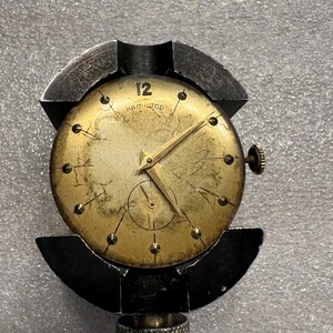 K&ouml;nnte beinhalten: Vintage Hamilton-Uhr mit goldfarbenem Zifferblatt, schwarzen Zeigern und einer kleinen Sekundenanzeige. Das Zifferblatt zeigt die Zahl 12 und den Markennamen. Die Uhr wird in einem schwarzen Metall-Uhrenhalter gehalten.