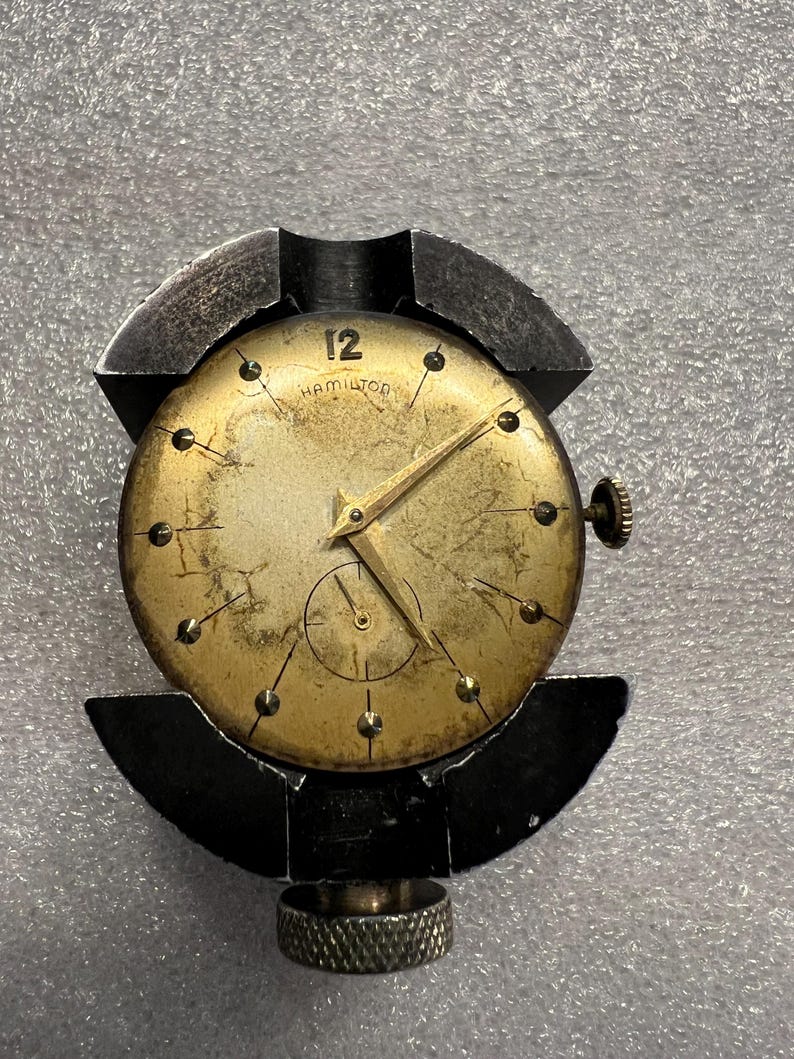 K&ouml;nnte beinhalten: Vintage Hamilton-Uhr mit goldfarbenem Zifferblatt, schwarzen Zeigern und einer kleinen Sekundenanzeige. Das Zifferblatt zeigt die Zahl 12 und den Markennamen. Die Uhr wird in einem schwarzen Metall-Uhrenhalter gehalten.