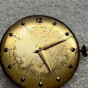 K&ouml;nnte beinhalten: Vintage Hamilton Zifferblatt mit goldfarbenem Finish. Das Zifferblatt hat schwarze Stundenmarkierungen, goldene Zeiger und die Zahl 12 oben. Das Wort "HAMILTON" ist unter der 12 aufgedruckt. Unten befindet sich ein kleines Zifferblatt.