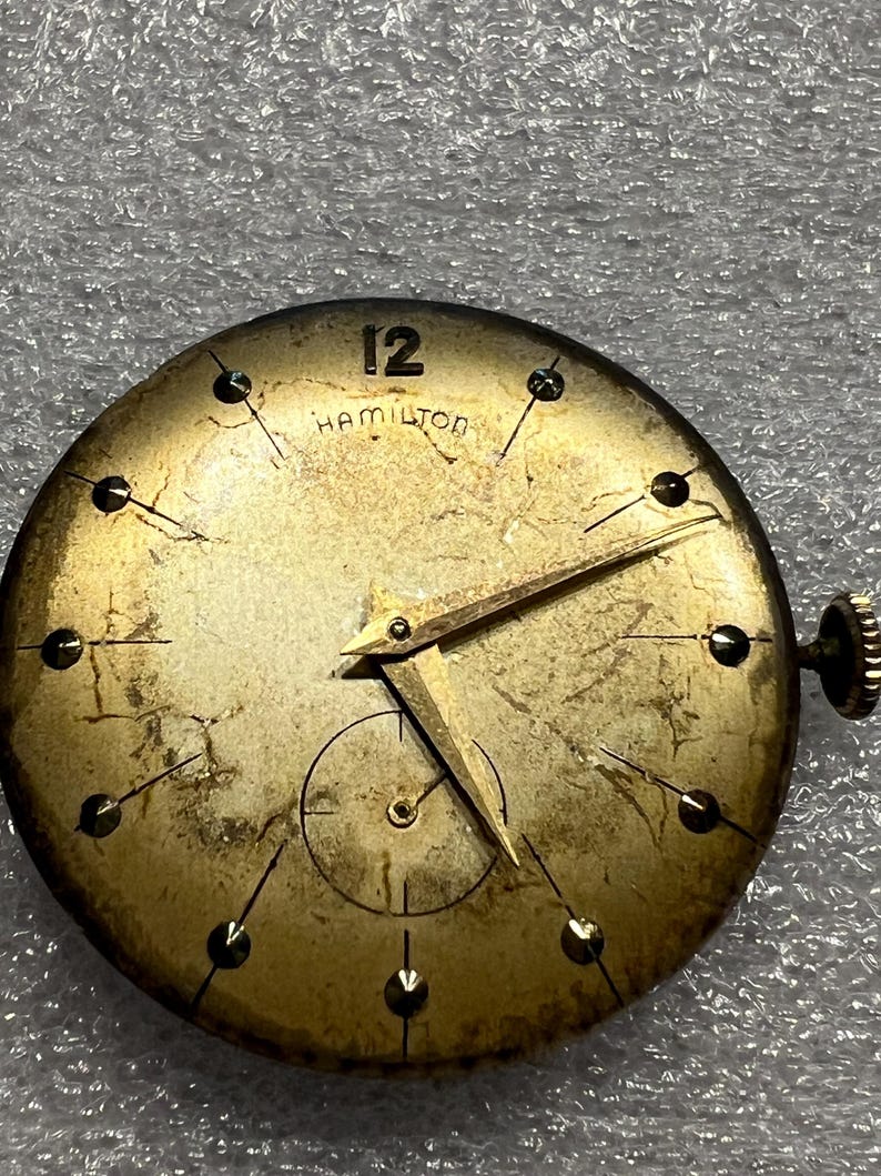 K&ouml;nnte beinhalten: Vintage Hamilton Zifferblatt mit goldfarbenem Finish. Das Zifferblatt hat schwarze Stundenmarkierungen, goldene Zeiger und die Zahl 12 oben. Das Wort "HAMILTON" ist unter der 12 aufgedruckt. Unten befindet sich ein kleines Zifferblatt.